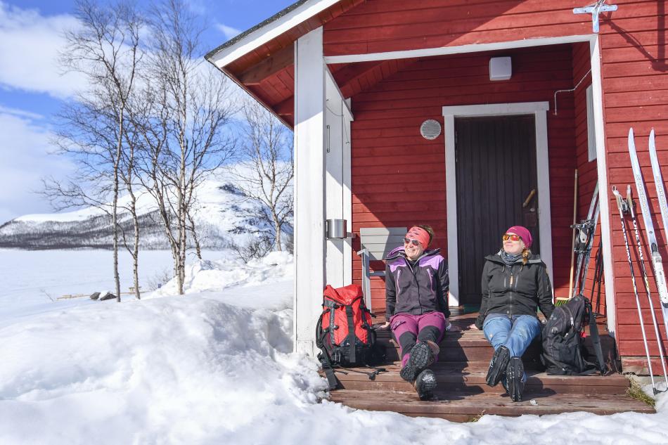 Two women are sitting in the sun on the steps of a small red cabin. They have their backbags and skis and poles out as well